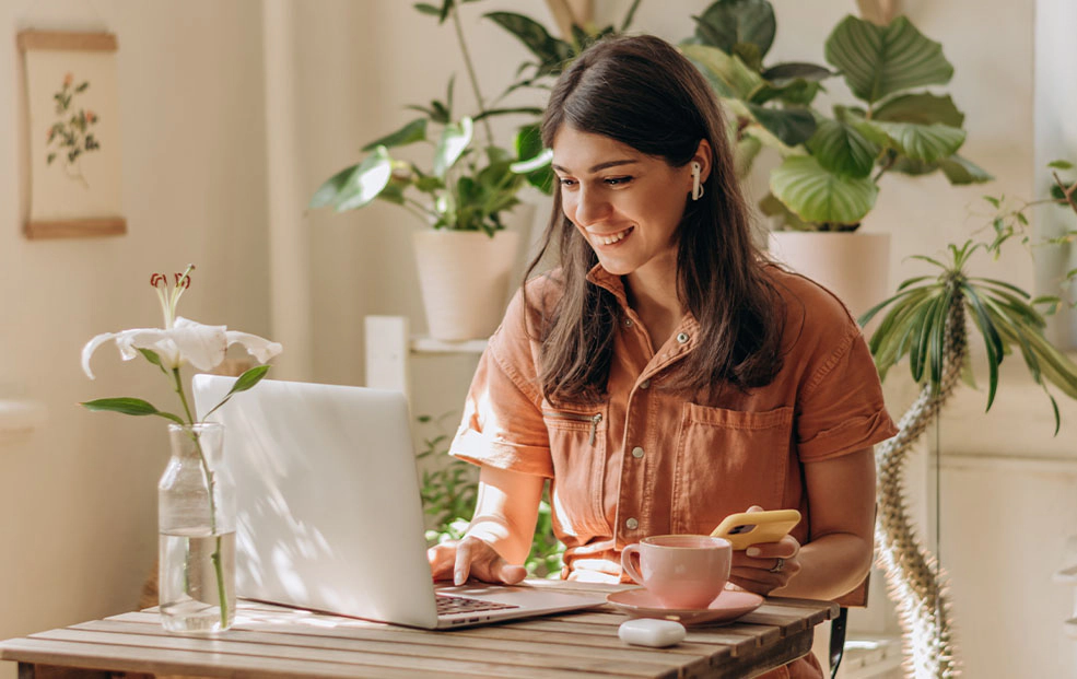 Smiling woman in an orange button-up top works on a laptop at a wooden table, with a coffee cup, a phone, and a white lily in a vase, surrounded by houseplants.