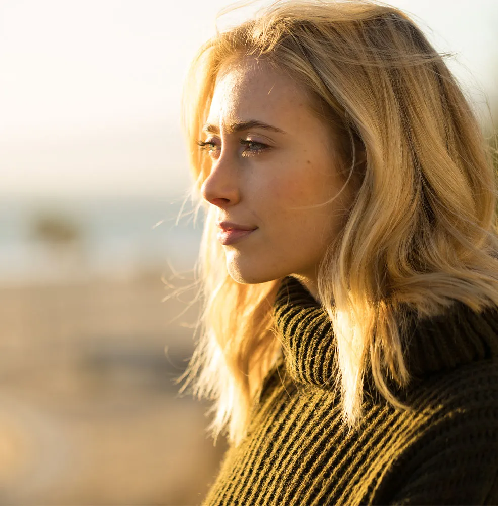 Woman with wavy blonde hair in a dark olive knit sweater gazing thoughtfully into the distance, lit by warm golden sunlight