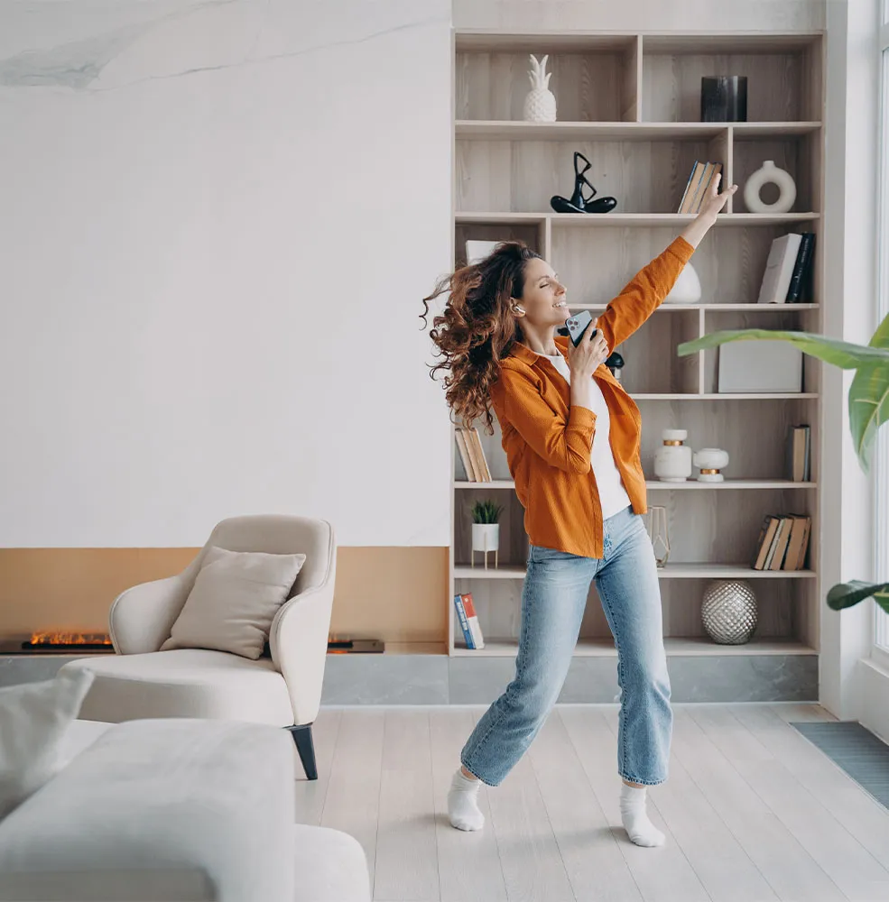 Woman in an orange shirt and jeans dancing joyfully in a modern living room, holding a phone like a microphone with one arm raised