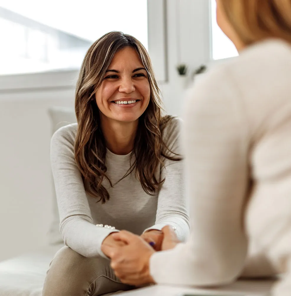 Smiling woman with long brown hair sitting on a couch and holding hands with another person in a bright, softly lit room